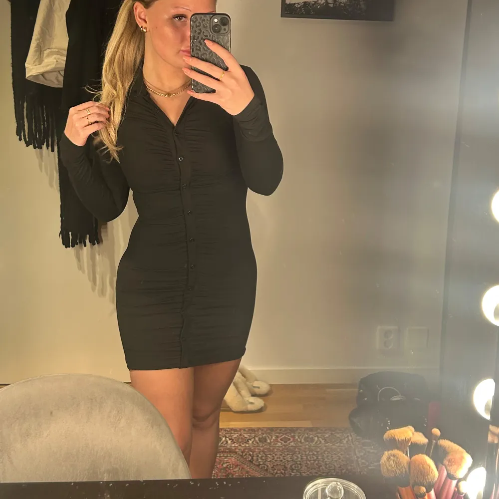 Snygg svart klänning med knappar framtill och långärmad design. Den har en tight passform som framhäver figuren och en kort längd som ger en modern look. Perfekt för en kväll ute eller en speciell tillställning.. Mekot.