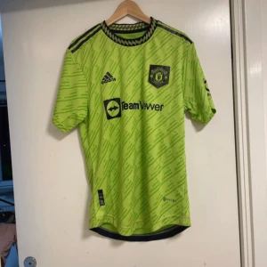 Grön fotbollströja från Adidas - Säljer en grön fotbollströja från Adidas med Manchester Uniteds emblem på bröstet. Tröjan har ett unikt mönster och korta ärmar. Perfekt för fans som vill sticka ut på läktaren eller planen.