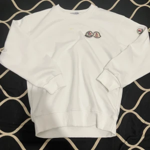 Moncler tröja - Säljer en stilren vit sweatshirt från Moncler med broderad logga på bröstet och ärmen. Tröjan har långa ärmar och ribbade muddar vid ärmslut och nederkant. Perfekt för en avslappnad look.