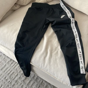 Svarta mjukisbyxor från Nike - Säljer ett par svarta mjukisbyxor från Nike med vita sidoremsor och logga. Byxorna har en elastisk midja och är perfekta för en avslappnad stil. De är tillverkade i ett mjukt och bekvämt material.