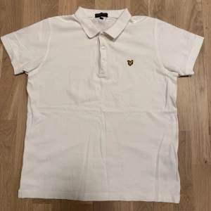 Vit pikétröja från Lyle & Scott - Säljer en klassisk vit pikétröja från Lyle & Scott med en broderad logga på bröstet. Tröjan har korta ärmar och en knappslå med tre knappar. Perfekt för en stilren och avslappnad look.