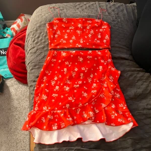 Röd blommig set med volang - Säljer en söt röd set med blommigt mönster och volangdetaljer. Settet har smala axelband och en omlottkjol som ger en fin rörelse. Perfekt för sommarens festligheter! 🌸S på linnet och M på kjolen 