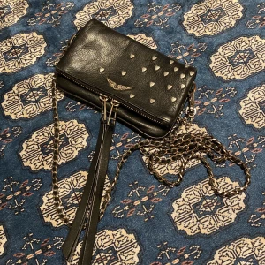 Svart axelväska med hjärtan från Zadig & Voltaire - Snygg svart axelväska i skinn från Zadig & Voltaire. Väskan har coola hjärtan som gör väskan speciell  och en kedja som axelrem. Perfekt för att ge din outfit en edgy touch. Den har en dragkedja för säker förvaring. Bra skick köpt på Plick. Kan inte riktigt måten men den är liten. Jämfört med en toffla för att se ungefär hur står den är💗