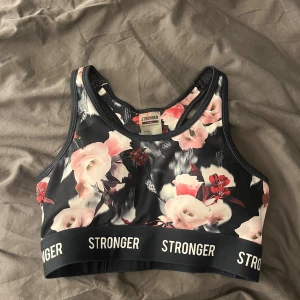 Blommig sporttopp från Stronger - Snygg sporttopp från Stronger med blommigt mönster i rosa och rött. Tillverkad av återvunnen polyester och elastan för en bekväm passform. Perfekt för träning med sitt elastiska band med logotypen 'STRONGER'.