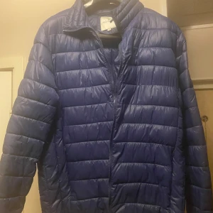 Blå quiltad jacka från Moncler - Säljer en snygg blå quiltad jacka från Moncler. Den är perfekt för kyligare dagar med sin pufferstil och långa ärmar. Jackan har en dragkedja framtill och är i nyskick. Den har också en cool detalj med ett märke på ärmen. Perfekt för både höst och vinter!