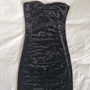 Svart axelbandslös klänning från Aft - Säljer en elegant svart axelbandslös klänning från Att. Den är gjord i ett sammetstyg som består av 95% polyester och 5% elastan, vilket ger en snygg, hudnära passform. Perfekt för festliga tillfällen! 