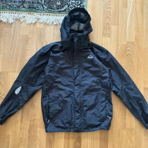 Svart vindjacka från McKinley - Säljer en svart vindjacka från McKinley, perfekt för utomhusaktiviteter. Jackan har en justerbar huva och dragkedja framtill. Den är märkt med 'Exodus 5000' på ärmen, vilket tyder på bra väderskydd. Passar perfekt för höst- och vårväder.