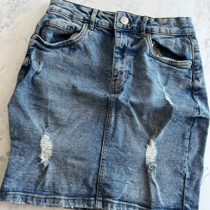 Blå jeanskjol med slitningar - Säljer en cool blå jeanskjol med slitningar framtill. Kjolen har en klassisk fyrficksdesign och stängs med knapp och dragkedja. Perfekt för en avslappnad vardagslook eller en utekväll med vänner. Passar bra till sneakers eller boots!