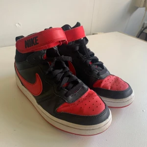 Svart och röd Nike sneakers - Säljer ett par coola Nike sneakers i svart och rött. De har snörning och en praktisk kardborreknäppning upptill. Skorna är i bra skick och perfekta för vardagsbruk eller träning. Sula med bra grepp och klassisk Nike-logga på sidan.