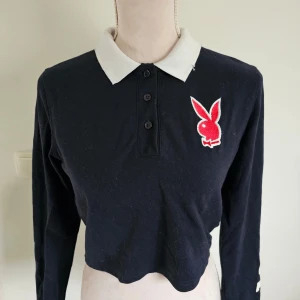 Svart piké crop top från Playboy x Missguided - Säljer en snygg svart pikétröja från Playboy x Missguided. Den har en vit krage och ett coolt Playboy-märke på bröstet. Tröjan är croppad och har långa ärmar med texten 'PLAYBOY' på ena ärmen. Kragen har en speciell design