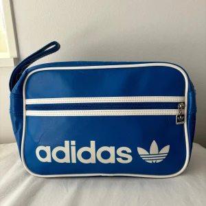 Blå handväska från Adidas - Säljer en snygg blå axelväska från Adidas med vit logga och detaljer. Väskan har en praktisk dragkedja och är perfekt för vardagsbruk. Den är i syntetmaterial och har en klassisk sportig stil.