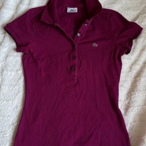 Vinröd pikétröja från Lacoste - Säljer en snygg vinröd pikétröja från Lacoste i storlek 36. Tröjan har korta ärmar och den klassiska krokodilloggan på bröstet. Perfekt för en casual look eller en dag på golfbanan. Knapparna framtill ger en stilren touch. Passar både till jeans och kjol!pris går att diskutera 
