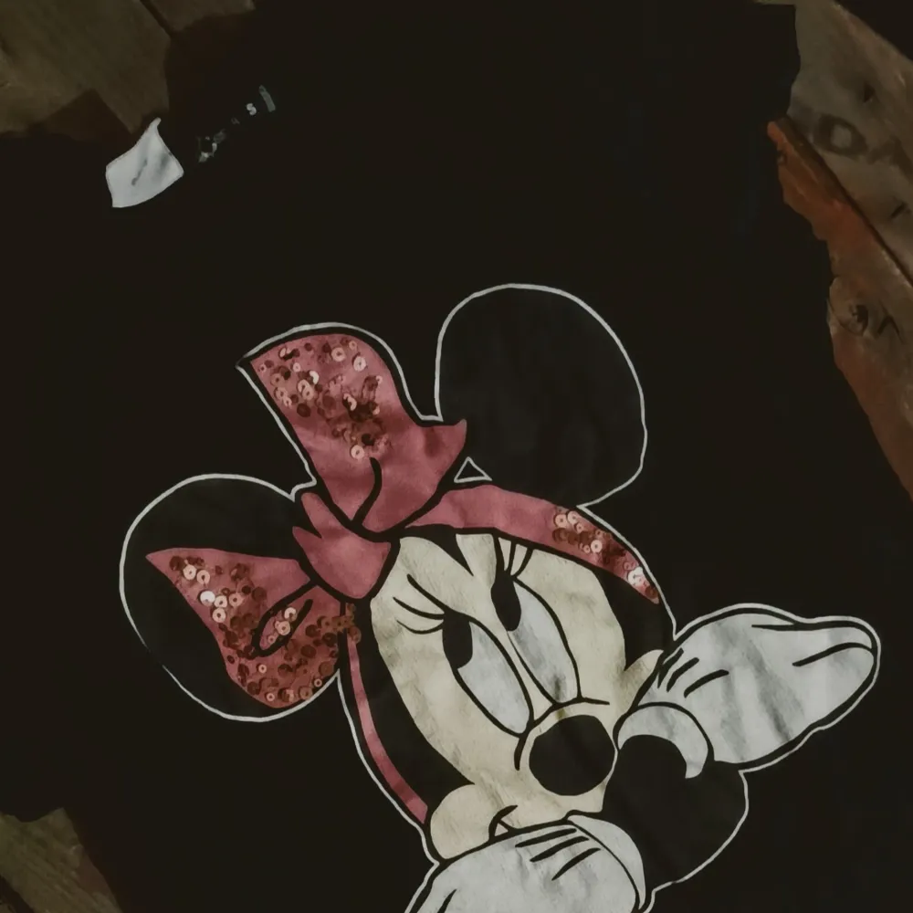 Säljer en svart t-shirt med ett stort tryck av Minnie Mouse på framsidan. Motivet har en söt rosa rosett med paljetter som ger en glittrig effekt. Perfekt för alla Disney-fans! 🖤. T-paidat.