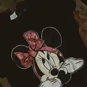 Säljer en svart t-shirt med ett stort tryck av Minnie Mouse på framsidan. Motivet har en söt rosa rosett med paljetter som ger en glittrig effekt. Perfekt för alla Disney-fans! 🖤