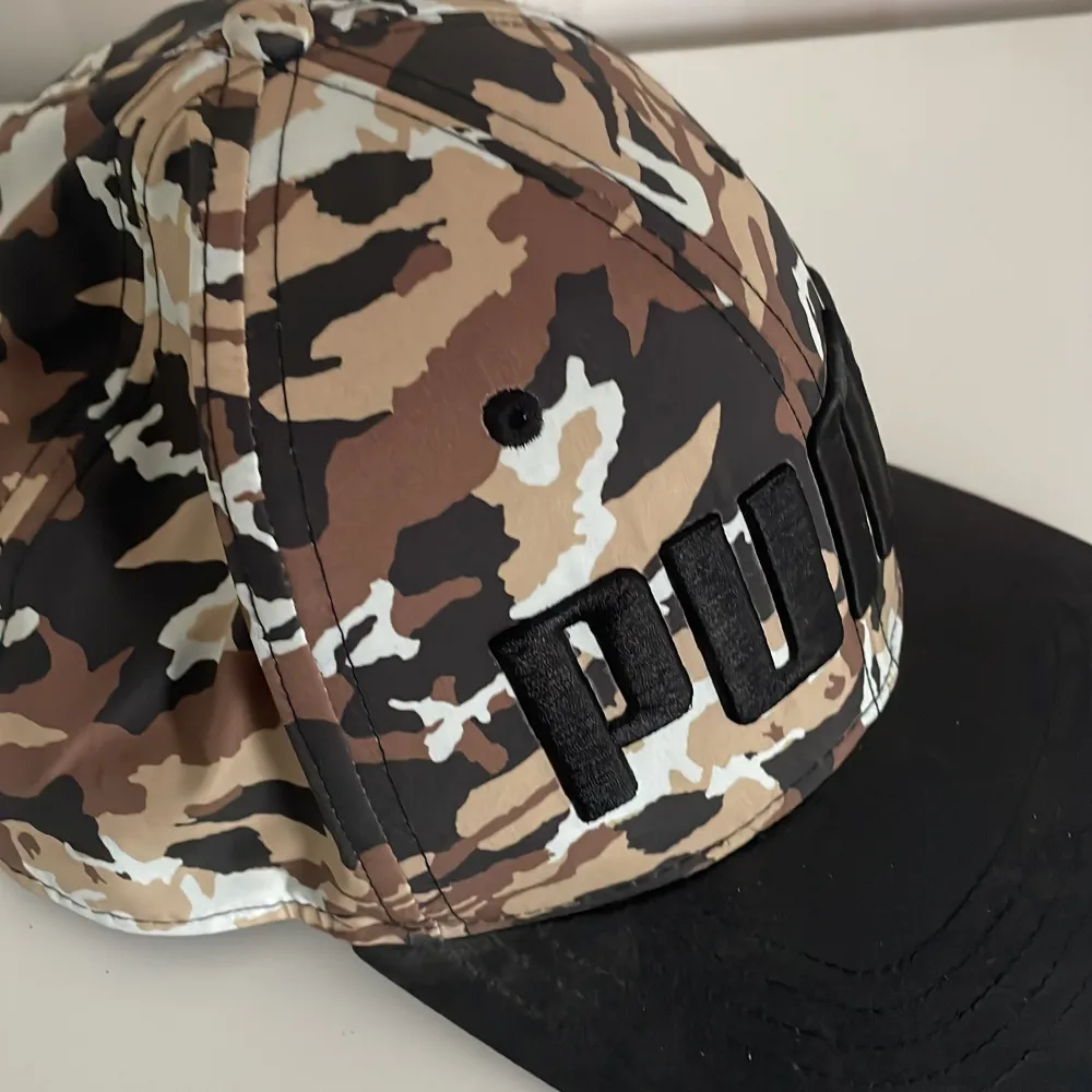 Säljer en snygg keps från Puma med ett coolt camouflage-mönster i bruna och svarta toner. Kepsen har en broderad Puma-logga framtill och en justerbar rem baktill för perfekt passform. Perfekt för både vardag och utomhusaktiviteter!. Asusteet.