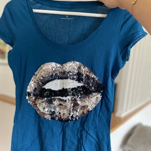 Blå t-shirt med paljettläppar - Säljer en cool blå t-shirt i bomull med ett stort paljettmotiv av läppar på framsidan. Den är kortärmad och perfekt för en avslappnad stil. Passar bra till både jeans och kjol. Perfekt för sommarens alla äventyr! 💙