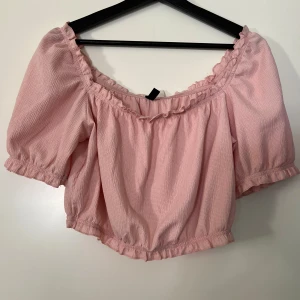 Rosa offshoulder topp från H&M Divided - Säljer en söt rosa offshoulder topp från H&M Divided. Den har korta ärmar och en fin volangdetalj runt halsringningen. Perfekt för sommardagar eller en avslappnad fest. Toppen är i ett lätt och luftigt material.