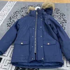 Mörkblå vinterjacka från Hampton Republic - Säljer en snygg mörkblå vinterjacka från Hampton Republic. Jackan har en mysig pälskantad huva och praktiska fickor med knappar. Den är perfekt för kalla dagar och har en cool logga på insidan. Dragkedjan är i guld och ger en extra touch av stil. Passar perfekt för vintern! ❄️