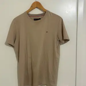 Säljer en snygg beige t-shirt från Tommy Hilfiger i bra skick. Den har en liten logga på bröstet och är gjord av mjuk bomull. Perfekt för en avslappnad stil och funkar året runt. Passar till både jeans och shorts!