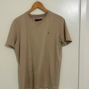 Beige t-shirt från Tommy Hilfiger - Säljer en snygg beige t-shirt från Tommy Hilfiger i bra skick. Den har en liten logga på bröstet och är gjord av mjuk bomull. Perfekt för en avslappnad stil och funkar året runt. Passar till både jeans och shorts!