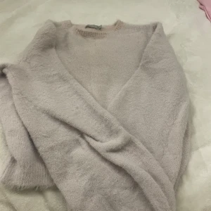 Beige fluffig tröja från Bershka - Mysig och fluffig beige tröja från Bershka i storlek XS. Perfekt för kyliga dagar med sin mjuka och varma känsla. Tröjan har en v-ringad design och långa ärmar, vilket gör den både stilren och bekväm. Passar perfekt till både vardag och mysiga hemmakvällar.