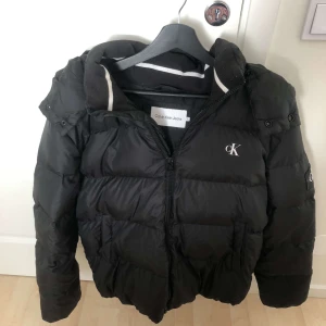 Svart pufferjacka från Calvin Klein - Säljer en snygg svart pufferjacka från Calvin Klein. Jackan har en dragkedja framtill och är quiltad för extra värme. Perfekt för kalla dagar! Den har en huva och långa ärmar, vilket gör den idealisk för vinter och höst. Passar både till vardags och lite finare tillfällen.