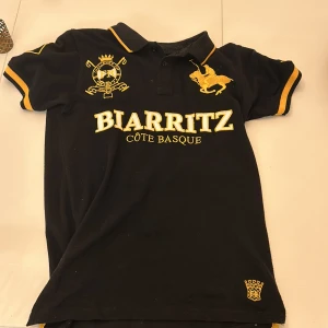 Svart pikétröja från Gravitee - Säljer en snygg svart pikétröja från Gravitee med gula detaljer och broderier. Texten 'BIARRITZ CÔTE BASQUE' pryder framsidan och 'POLO TEAM 1890' finns på baksidan. Perfekt för en avslappnad men stilren look. Passar bra till både vardag och fest!