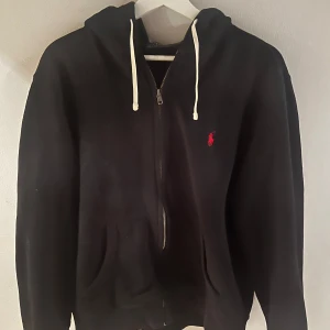 Svart hoodie från Polo Ralph Lauren - Säljer en snygg svart hoodie från Polo Ralph Lauren i storlek M. Den har en dragkedja framtill och en klassisk logga i rött på bröstet. Perfekt för höst och vinter, med en skön passform och huva med snören. Passar både till vardags och chill.