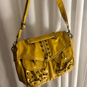 Gul axelväska med nitar - Säljer en cool gul axelväska med nitar och dragkedja. Väskan har två fickor framtill och justerbar axelrem. Perfekt för att ge din outfit en edgy touch! 🖤