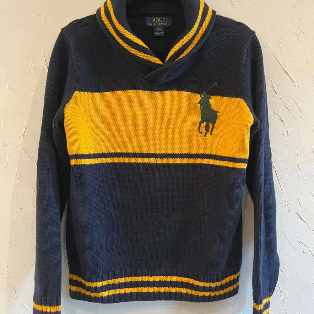 Säljer en snygg mörkblå tröja från Polo Ralph Lauren med en bred gul rand över bröstet och det klassiska logotypen. Tröjan har långa ärmar och en skön krage. Perfekt för höst och vinter!. Tröjor & Koftor.