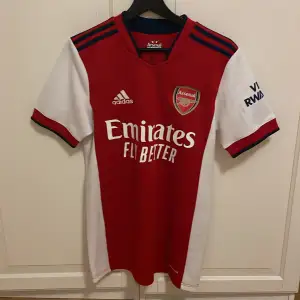 Säljer en snygg röd Arsenal fotbollströja från Adidas i mycket bra skick. Den har Arsenal-loggan och 'Emirates Fly Better' tryckt på framsidan. På baksidan står det 'Ødegaard' och nummer 8. Perfekt för alla Arsenal-fans! ⚽️Tröjan är storlek S men passar M galant