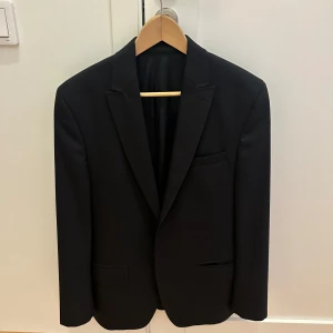 Svart kavaj från Riley - Snygg svart kavaj från Riley i storlek 48. Perfekt för både formella och semi-formella tillfällen. Kavajen har en klassisk design med två knappar och stilrena detaljer i rött på insidan. Passar utmärkt för höst och vinter. Använd den på festen eller balen för en stilren look.