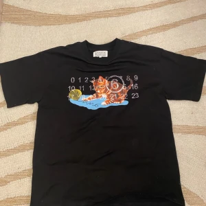 Maison Margiela tshirt - Säljer en svart t-shirt från Maison Margiela med en gullig katt på framsidan. Perfekt för en avslappnad stil eller att bära till vardags. Storleken är S till M