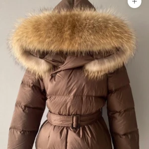 Brun dunjacka med pälsdetalj - Säljer en helt ny supermysig brun dunjacka med stor fluffig päls på huvan. Jackan har ett bälte i midjan för en snygg passform och långa ärmar som håller dig varm hela vintern. Perfekt för kalla dagar när du vill vara både stilren och varm!