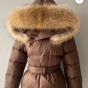 Brun dunjacka med pälsdetalj - Säljer min helt nya supermysig brun dunjacka med stor fluffig päls på huvan. Jackan har ett bälte i midjan för en snygg passform och är perfekt för kalla vinterdagar. Den är långärmad och ser riktigt varm ut. Perfekt för både vardag och festliga vinterkvällar!