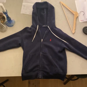Mörkblå hoodie från Ralph Lauren - Säljer en snygg mörkblå hoodie från Ralph Lauren i storlek XS. Den har en dragkedja framtill och en klassisk röd logga på bröstet. Perfekt för höst och vår, med långa ärmar och en skön huva. Passar både till vardags och chillkvällar.  Den är i topp skick