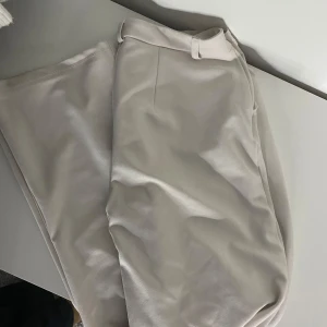 Beige kostymbyxor - Säljer ett par stilrena beige kostymbyxor i mycket bra skick. De har en klassisk design med knappar och bälteshällor, perfekt för både kontoret och finare tillfällen. Materialet känns mjukt och bekvämt, vilket gör dem sköna att bära hela dagen.