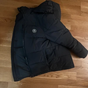 Svart pufferjacka från trapstar  - Säljer en svart pufferjacka från Trapstar i bra skick. Den har en stor huva och dragkedja framtill. Perfekt för kalla dagar med sin quiltade design. Jackan har långa ärmar och en cool logga på ryggen. Passar perfekt för vintern och hösten! Pris kan även diskuteras 