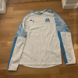 Vit och blå långärmad windbreaker från Marseille  - Säljer en snygg vit och blå långärmad windbreaker från Marseille . Tröjan har ett Marseille-emblem på bröstet och coola blå mönstrade ärmar. Perfekt för träning eller att heja på ditt lag! 🏆