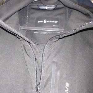 Svart jacka från Sail Racing - Säljer en svart jacka från Sail Racing i storlek medium. Den har en stilren design med dragkedja och en diskret logga på bröstet. Perfekt för höst och vår med sin funktionella och snygga look. Jackan är i mycket bra skick och redo för nya äventyr.