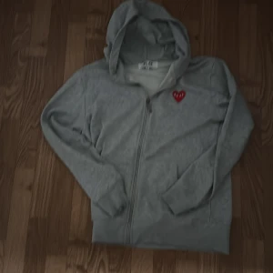 Grå hoodie med hjärtdetalj - Mysig grå hoodie med dragkedja och en söt röd hjärtdetalj på bröstet. Perfekt för kyliga dagar eller mysiga hemmakvällar. Den har långa ärmar och en praktisk huva.