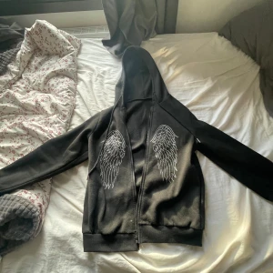 Svart hoodie med vingdetaljer från Shein - Säljer en svart hoodie från Shein i storlek XS. Den har snygga vingdetaljer i strass på framsidan och en dragkedja. Materialet är mjukt med 95% polyester och 5% elastan.