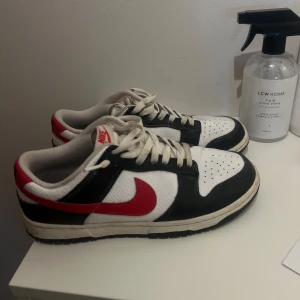 Nike Dunk sneakers i svart, vitt och rött - Säljer ett par Nike Dunk sneakers i klassisk färgkombination av svart, vitt och rött. Skorna har en snygg design med snörning och är i bra skick. Perfekta för både vardag och festliga tillfällen. Storlek är inte angiven, men de ser ut att vara i standard herrstorlek. Rengörs innan köp 