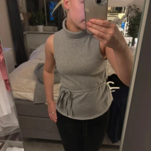 Grå ärmlös polotopp från Zara - Säljer en stilren grå ärmlös polotopp från Zara i storlek S. Toppen har en snygg knytdetalj i midjan som ger en fin siluett. Perfekt för både vardag och festliga tillfällen. Materialet är mjukt och bekvämt.