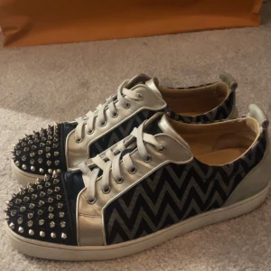 Christian Louboutin sneakers med nitar - Säljer ett par feta Christian Louboutin sneakers i mycket bra skick! De har ett blått och silver zigzag-mönster med snygga nitar på toecapen Skorna har den klassiska röda sulan och är perfekta för att sticka till festen. Storlek 43-44