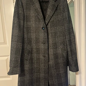 Rutig kappa från Sand - Säljer en elegant rutig kappa från Sand i svart och grått. Den har en klassisk design med knappar framtill och är perfekt för höst och vinter. Materialet är en blandning av viskos och syntet, vilket ger en skön känsla. Kappan är i nyskick aldrig använd  och passar perfekt för både vardag och festliga tillfällen. Den är i storlek 50 som medium large nypris 4499