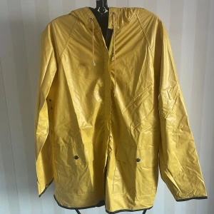 Gul regnjacka - Säljer en snygg gul regnjacka i bra skick. Den har långa ärmar och praktiska fickor med knappar. Perfekt för regniga dagar och håller dig torr och stilren. Jackan har en klassisk design med en mörk insida och detaljer i form av knappar och dragsko vid huvan. Vid armslutet är det ett litet hål, i övrigt som ny.