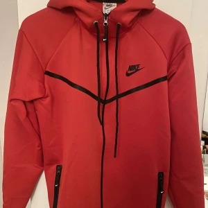 Röd hoodie från Nike - Säljer en snygg röd hoodie från Nike i mycket bra skick. Den har en dragkedja framtill och en justerbar huva med svarta snören. Perfekt för höst och vår, med långa ärmar och en sportig look. Svarta detaljer ger en cool kontrast. Passar perfekt för träning eller vardagsbruk!