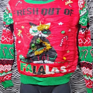 Jultröja med kattmotiv - Säljer en rolig och färgglad jultröja med ett kattmotiv och texten 'Fresh Out of Falalas'. Tröjan är röd med gröna och vita detaljer samt dekorerad med julgranskulor och glitter. Perfekt för julfesten eller som en kul present! 🎄