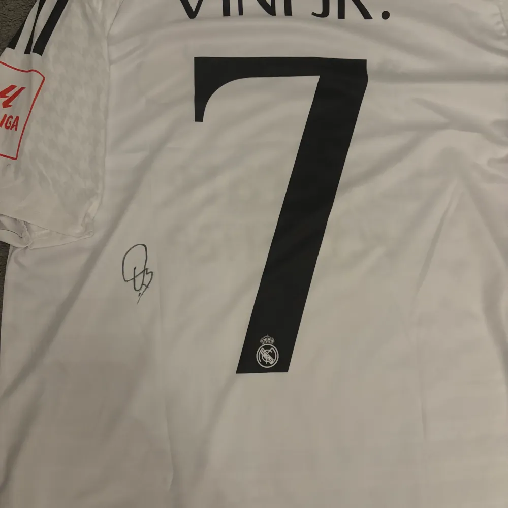 Säljer en vit Real Madrid fotbollströja från Adidas i mycket bra skick. Tröjan har korta ärmar och är dekorerad med klubbens emblem och sponsorlogga. På baksidan finns namnet 'VINI JR.' och nummer 7. Perfekt för alla fans av laget!. T-shirts.